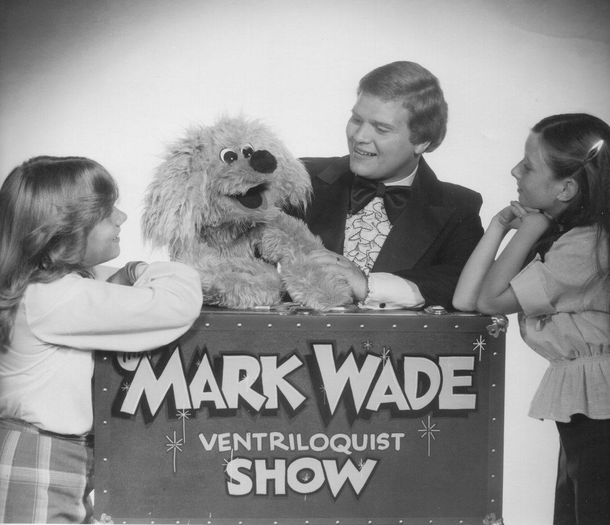 A young Mark Wade in a tuxedo with a dog figure and two children, leaning on a trunk lettered The Mark Wade Ventriloquist Show